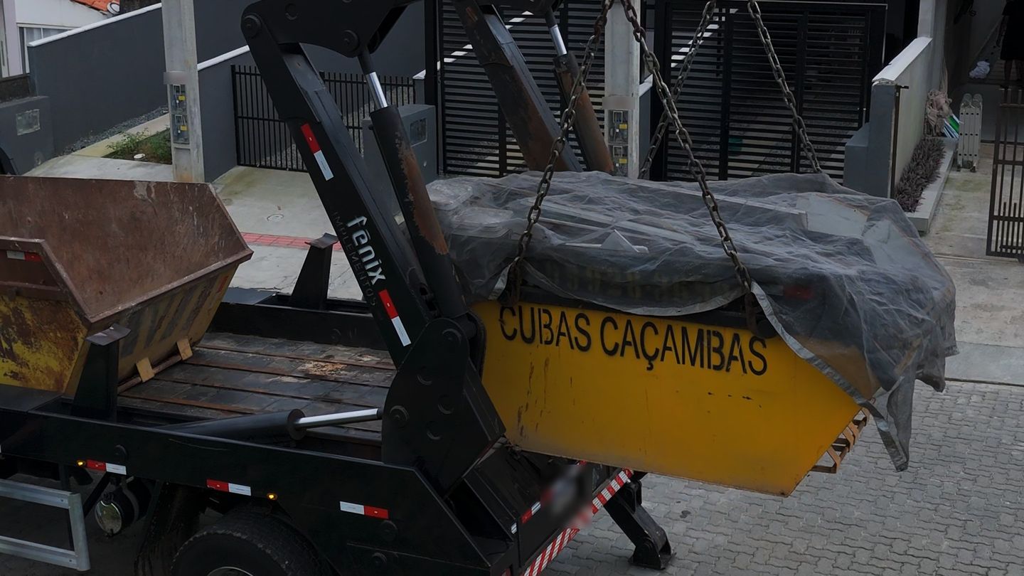 Caminhão da Cubas Caçambas içando uma caçamba amarela coberta com lona, com o nome da empresa na lateral, em frente a um sobrado