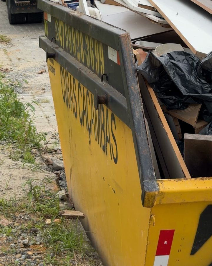 Caçamba amarela da Cubas Caçambas cheia de entulho de reforma, com tábuas, baldes e sacos, na beira da rua
